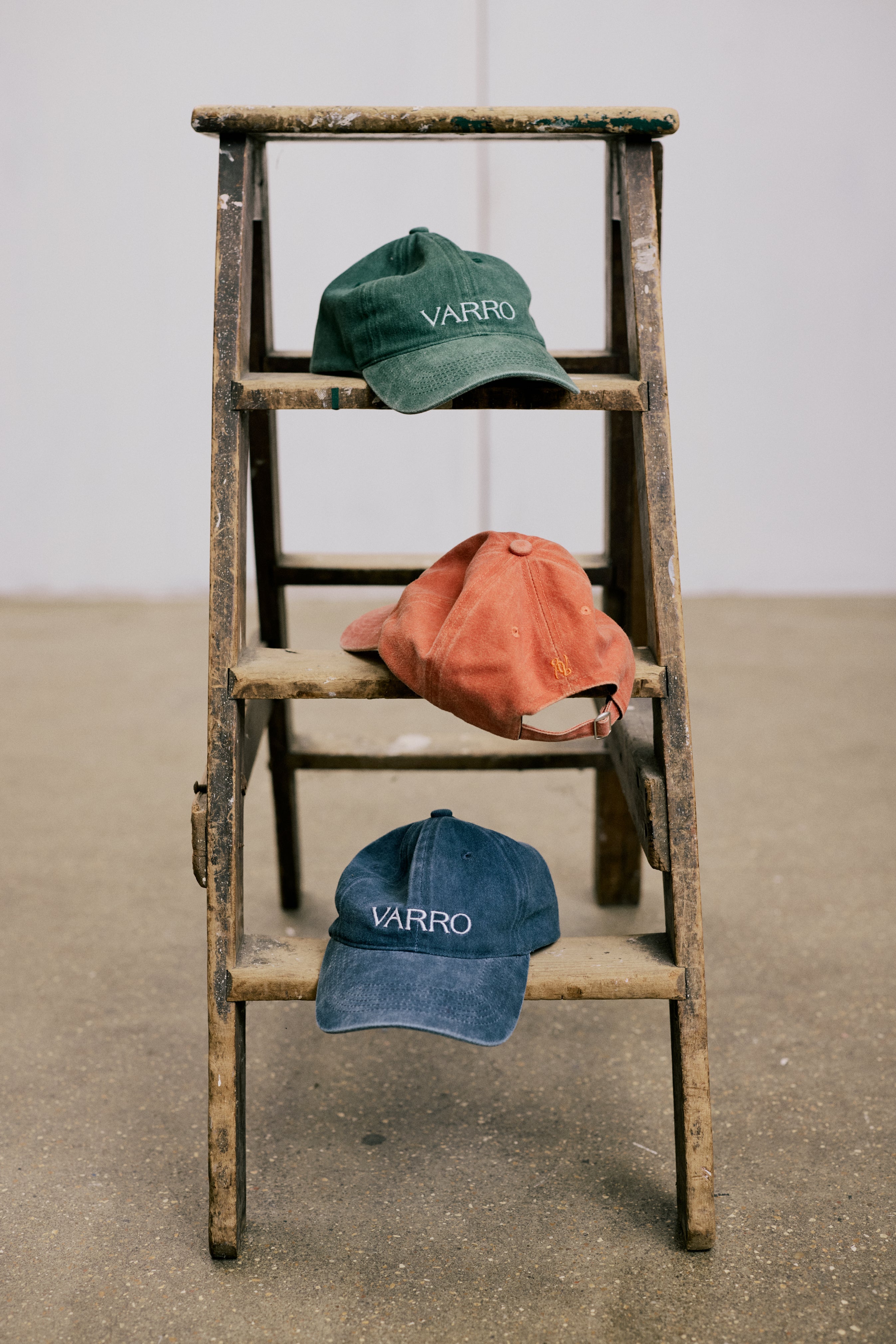 Stacked trio of VARRO vintage-washed cotton caps in forest green, midnight navy, and ember orange, showcasing embroidered logos and curved brims