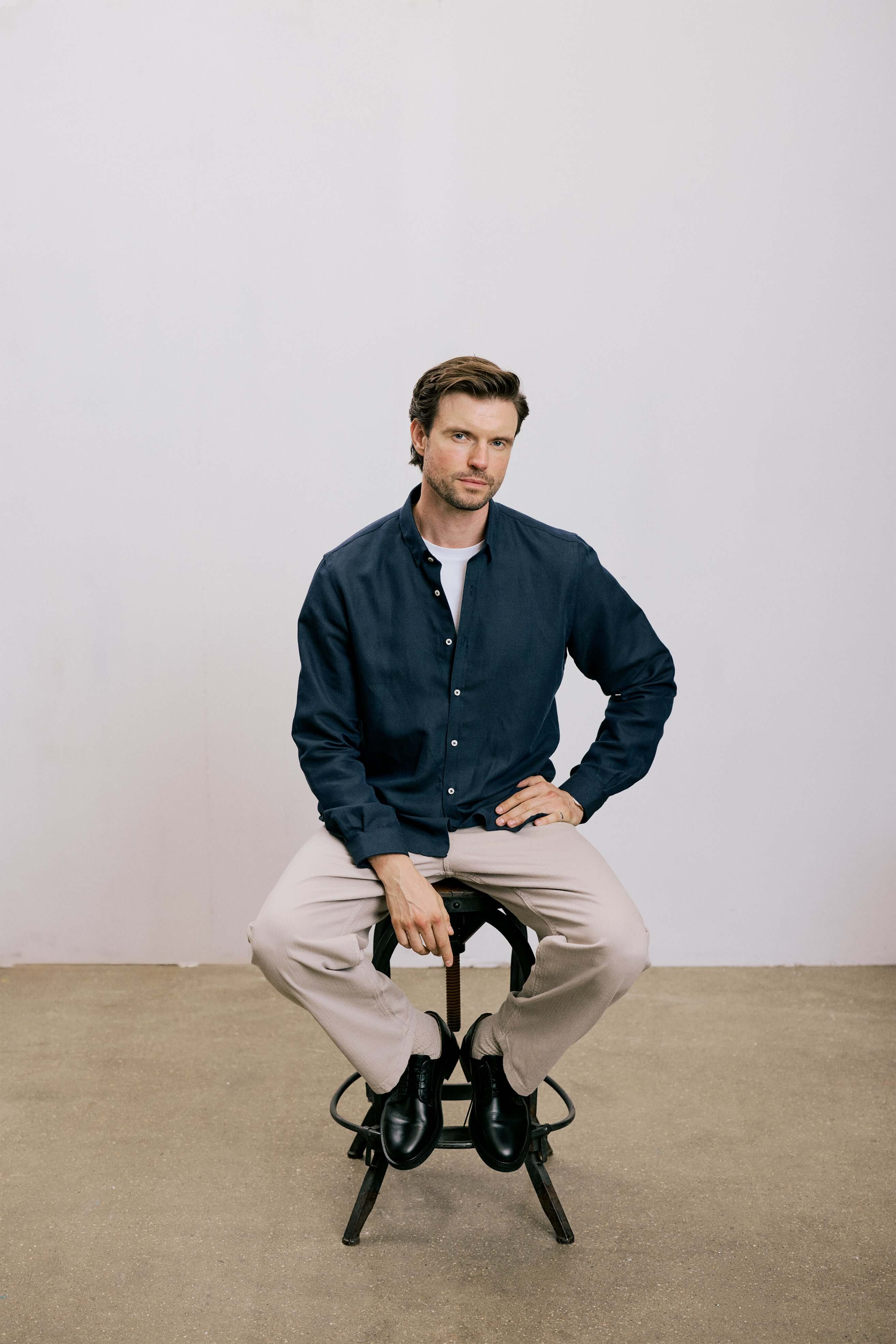 Model wearing VARRO Evelyn Midnight Navy shirt with hidden button-down collar, relaxed fit, seated on stool.